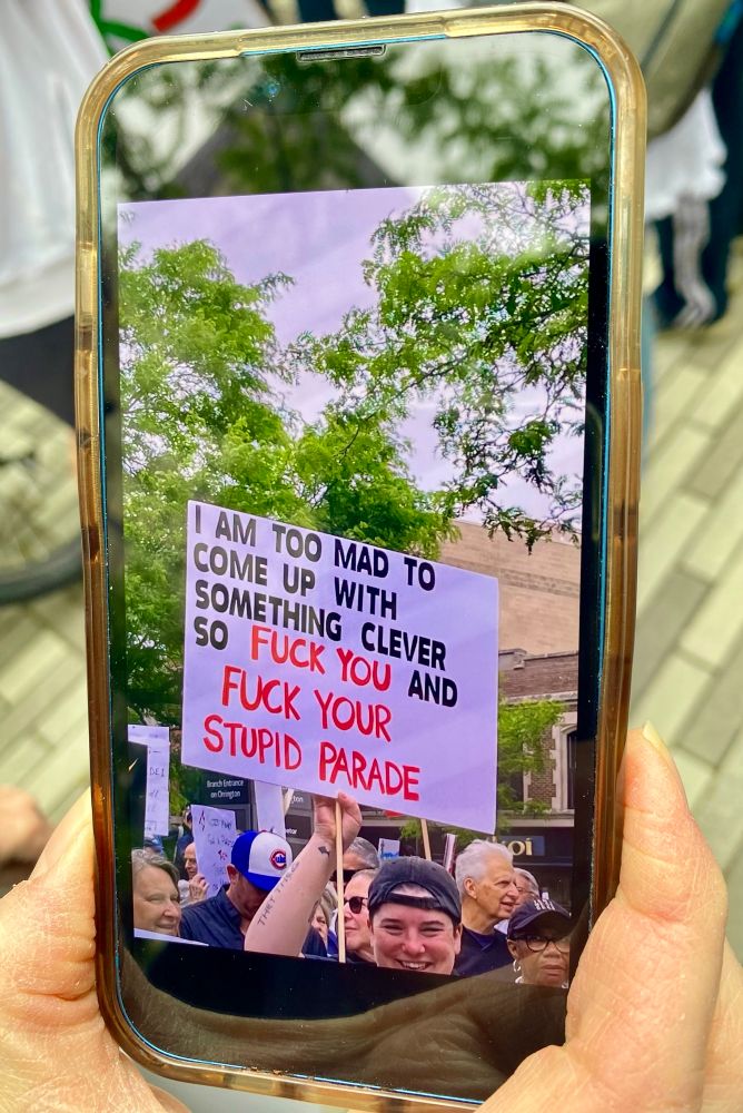 A smiling, twenty-something years old, Caucasian man wearing a backwards baseball cap holds up a sign that reads, “I am too mad to come up with something clever so fuck you and fuck your stupid  parade,”