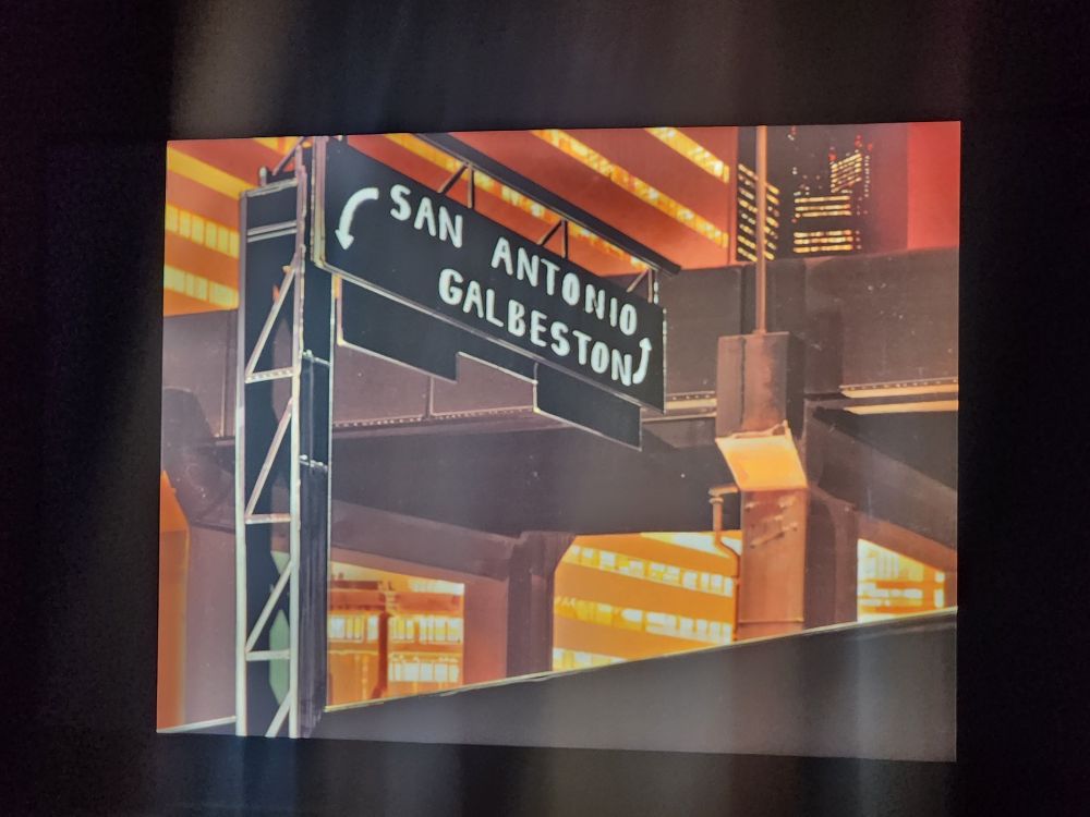 A screenshot from Bubblegum Crisis. It's a street sign over a highway that says "San Antonio" and an arrow to the left and "Galbeston" with an arrow to the right.