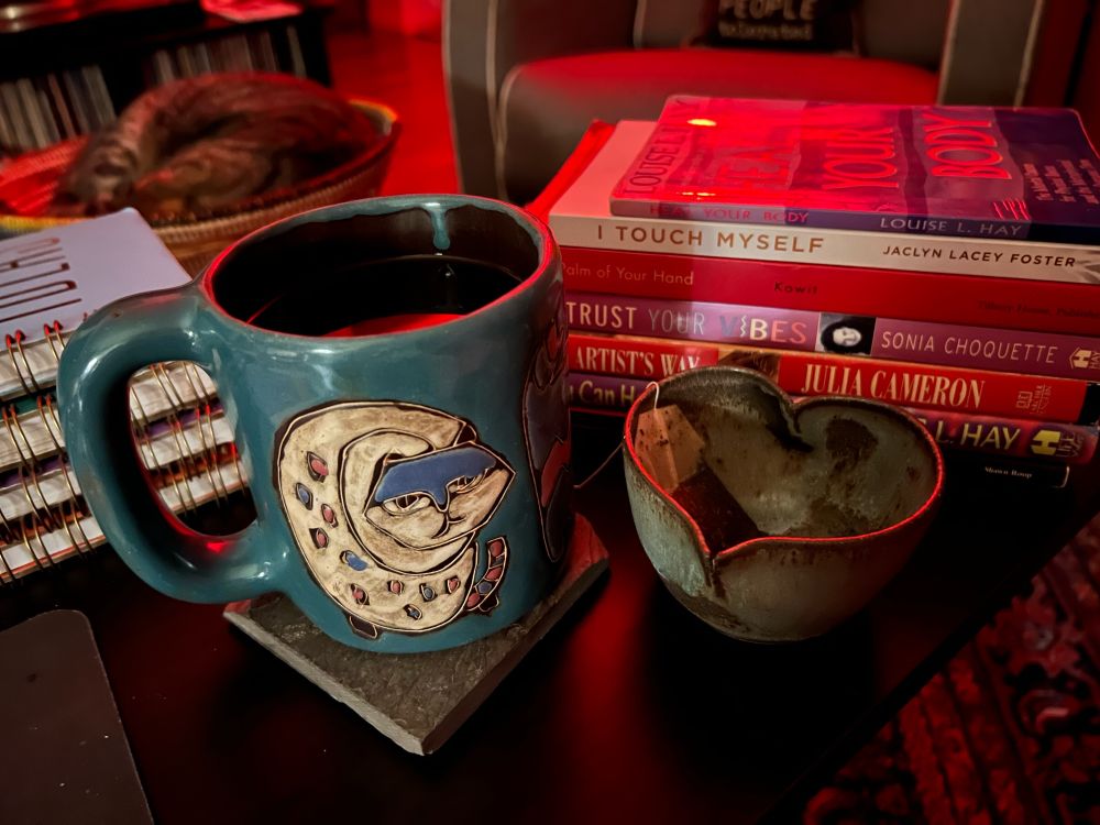 A sleeping cat is curled up in a basket on a coffee table in the top left corner. Spiral notebooks, and various reading material are stacked in the mid ground; a ceramic mug decorated with stylized cats is filled with peppermint tea sits on a stone drink coaster, while a heart shaped pinch pot holds the spent tea bag beside it.