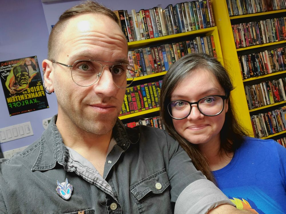 This user - Steel A Jeeg - and user Alykat717 photographed in front of a display of colorful VHS tapes, the tapes feature names indicating many of them are grindhouse and x-rated films from the 70s and 80s