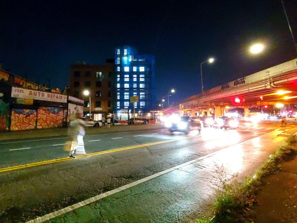 A stylized photograph of a city street showing a tall building under constructions at the center, a busy underpass on the right, and a graffiti covered auto shop on the left