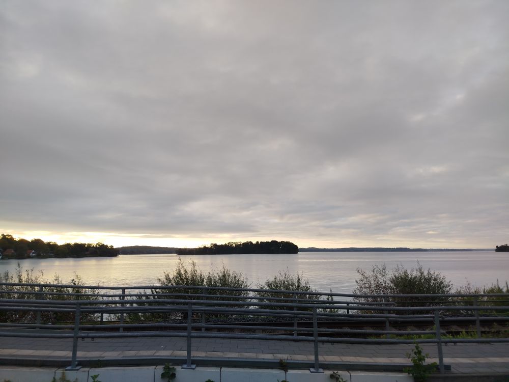 Sonnenaufgang am Plöner See. Im Vordergrund Geländer, dahinter Uferböschung und dann der See 