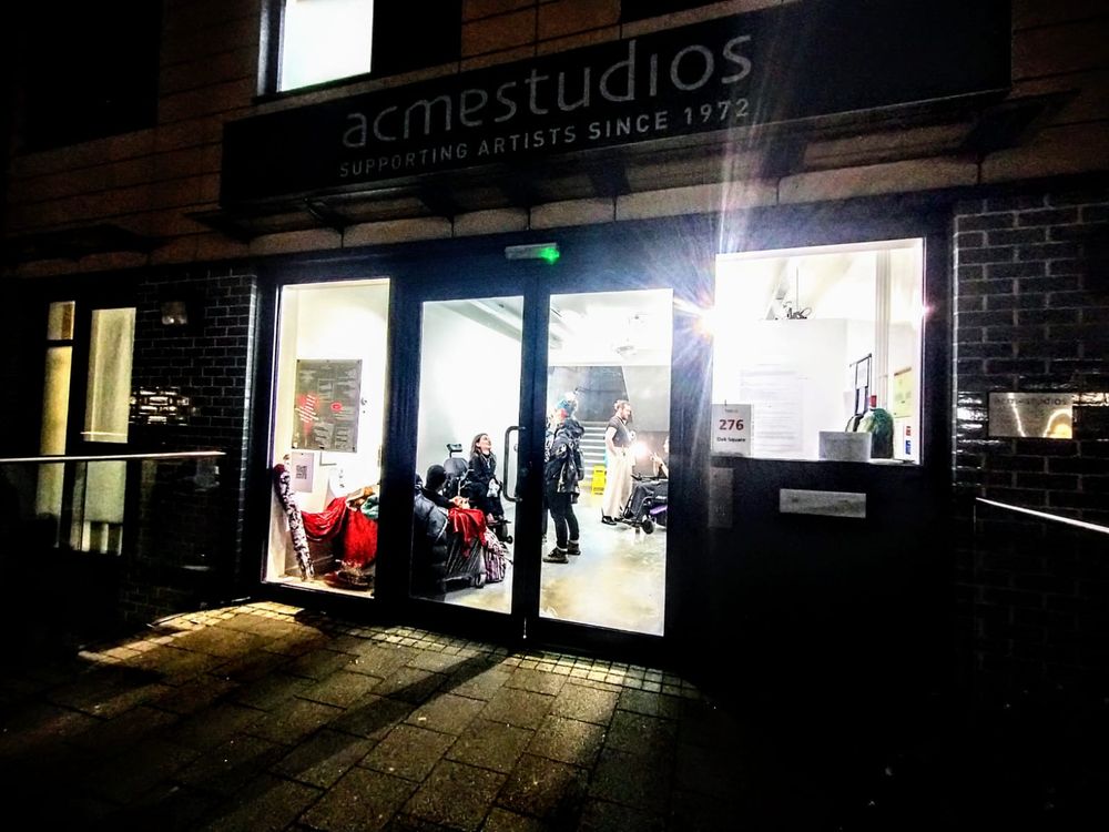 ACME Studios since 1972, sign above 4 windows with lights on and a dozen or so people inside. Some sat, stood and two using wheelchairs. A red & black poster in the window and red velvent on the seats. Its dark outside. 