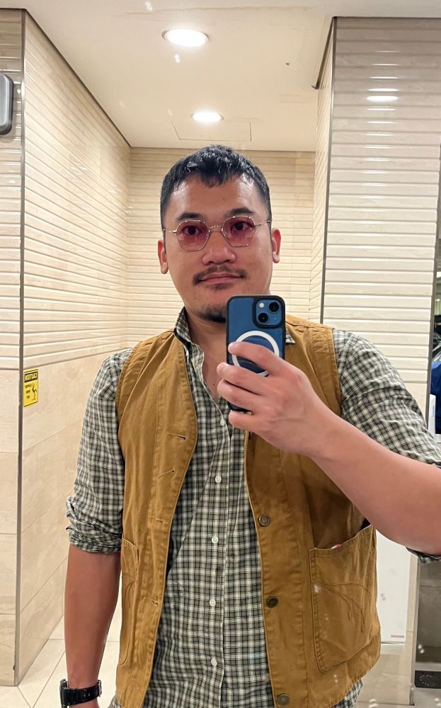 Selfie of a guy wearing pink glasses, a green plaid shirt and a brown vest.