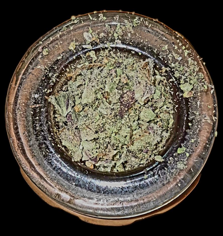 Cannabis Flower Grinds
A high-contrast, overhead macro shot of finely ground cannabis flower in a clear glass container. The ground material is a mix of deep greens and hints of purple and brown, showing a good bowl of potent flower. The glass container has a thick, glossy rim, and the black background emphasizes the texture and detail of the plant material.