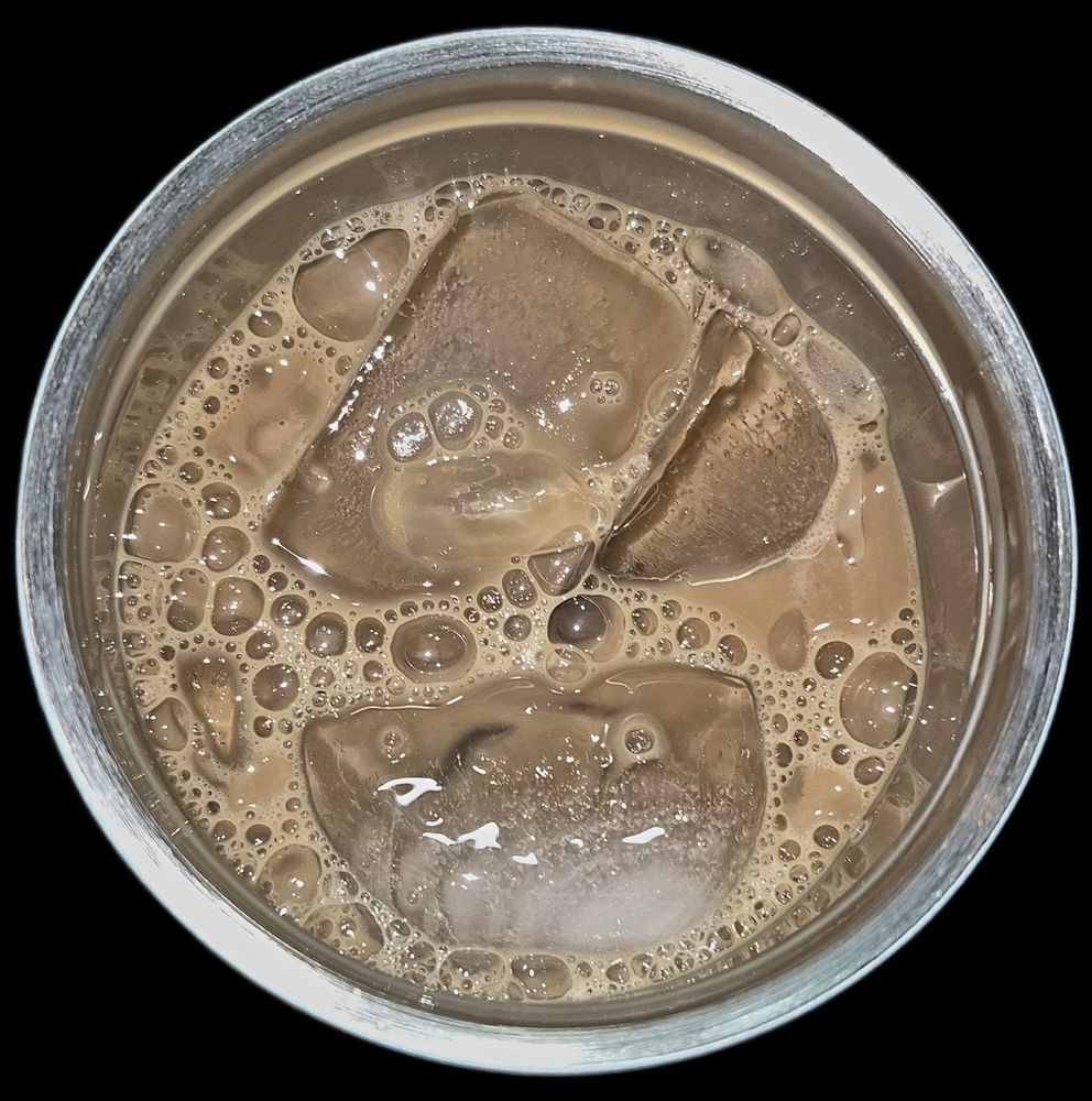 A tightly cropped, overhead view of a creamy vanilla iced coffee in a clear metal-rimmed glass or tumbler, set against a black background. The liquid is a light, milky tan color, with two large, glistening ice cubes partially submerged. The surface is covered in a delicate layer of silky, uniform light-brown foam and shiny bubbles that cling to the ice, creating a rich texture above the cold, refreshing drink.