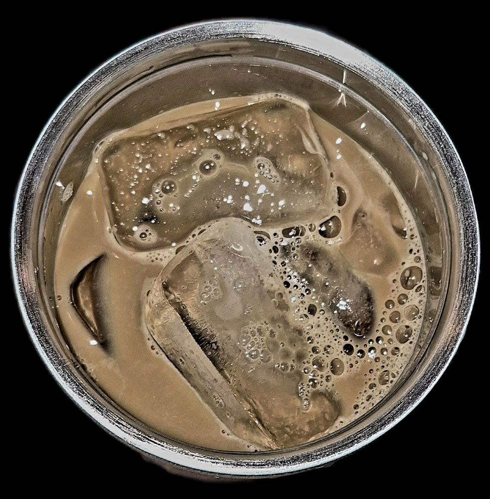 A dramatic, top-down view of an iced coffee or milk-based beverage in a metal-rimmed cup. The liquid is a light, creamy brown color. Dominating the frame are two large, semi-submerged, clear ice cubes with visible cracks and texture. The surface of the drink is covered in a multitude of tiny bubbles and foam, particularly concentrated around the edges of the ice. The background is a stark, solid black, making the reflective surface of the drink and ice the central focus.