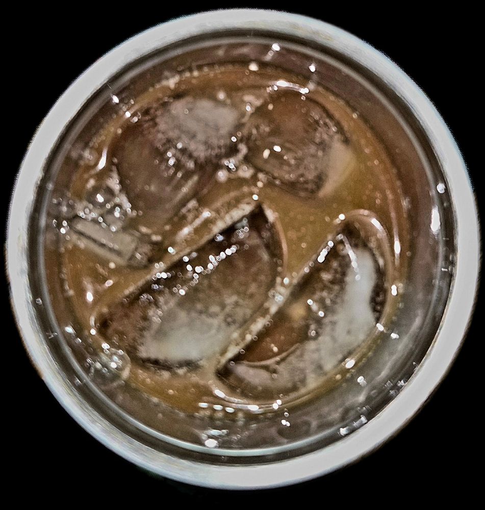 A top-down, high-contrast close-up looking into a cup of rich, deep mahogany cold brew coffee. Large, irregular ice cubes float semi-submerged in the dark liquid, their surfaces glistening with melting water and showing internal crystalline fractures. Clusters of tiny bubbles cling to the edges of the ice and the liquid’s surface, catching the light. Sharp, bright white reflections dance across the wet ice and the glossy, oily surface of the coffee, all framed by a soft-focus white rim against a stark black background.