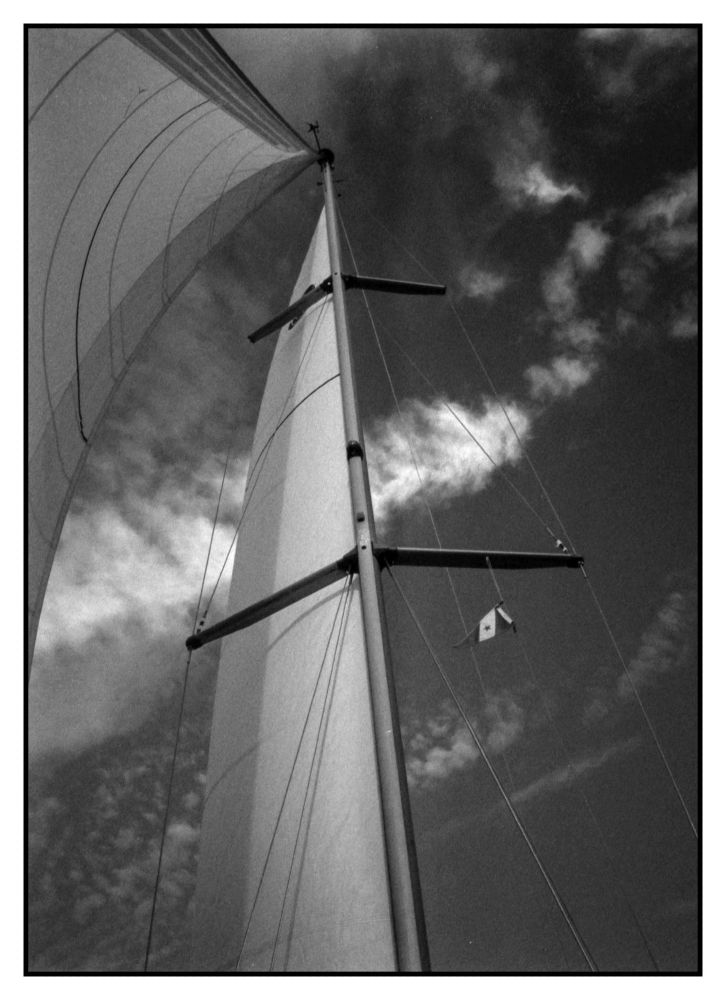 Looking up at the mast from the bow with the main and jib full and a bit of a stripe of cloud in the clearish sky.