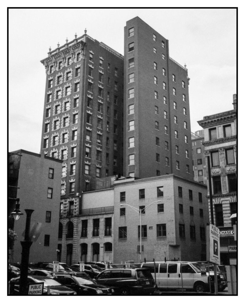 A group of buildings rising up above the parking lot, old structures different shapes and the striking one to me of the tall narrow looking one right of center. 