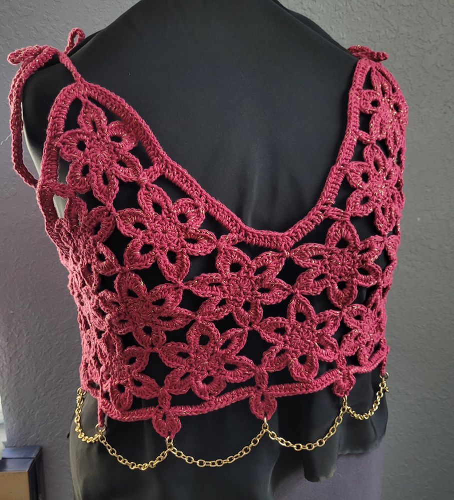 A crocheted top is shown on a mannequin. It is made of many 6 pointed flowers attached together. The flowers are made of pink yarn with gold thread mixed in for sparkle. At the bottom are individual petals with gold chain draped through them as an accent. 