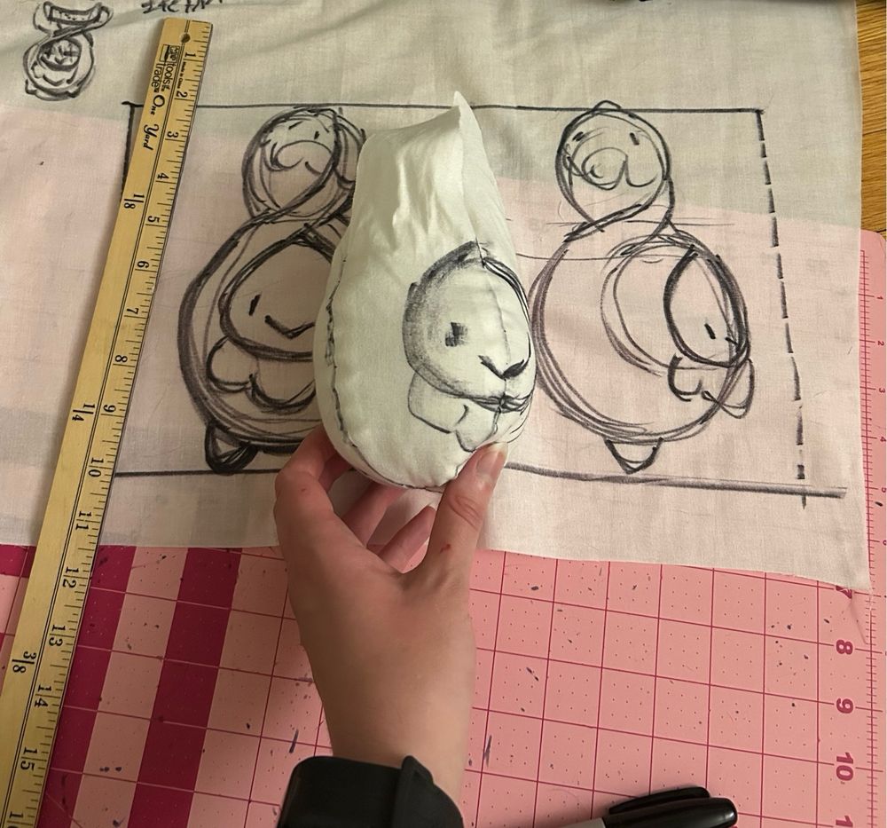 the beginnings of a prototype plush is being held in front of the blueprint sketches. its sized properly, currently only consisting of a body with some loose fabric bits atop its head in leu of its buds.
