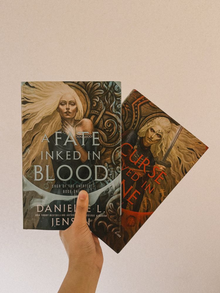 Holding up a copy of Danielle L. Jensen’s A Fate Inked in Blood and behind it, a copy of A Curse Carved in Bone. 