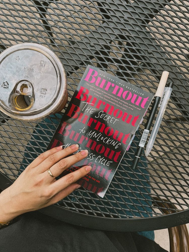 A cup of iced coffee, the book Burnout by Emily and Amelia Nagoski, and two pens on a patio table