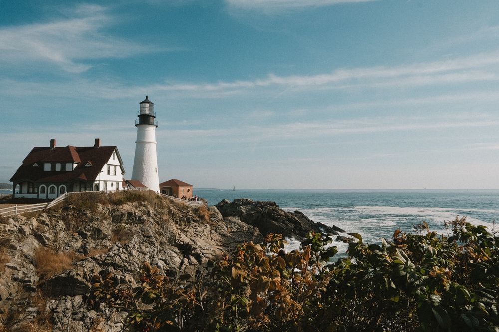 A house and a lighthouse on a rocky shoreline with the ocean behind it and foliage in the foreground 