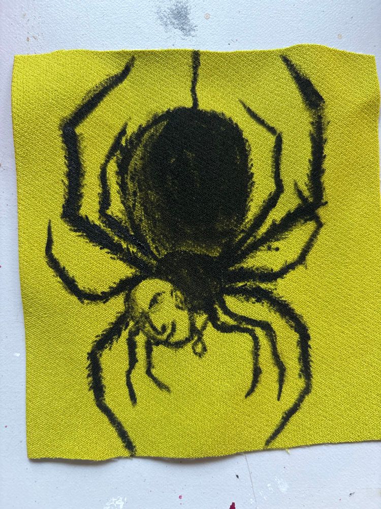 Green piece of fabric with a spider with a bald human head wearing a locket painted on it in black passing