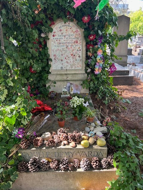 in august 2025 the white marble gravestone of jacques demy and agnes avarda is surrounded by green vines, interwoven with fake flowers, and a string of prayer flags reaches out from the top to the right edge of the image. the gravestone is covered with red lipstick marks and doodles. the grave in front is covered with dead flowers, pine cones, and potatoes, a reference to varda's last good film that didn't involve jr, les glaneurs et la glaneuse.
