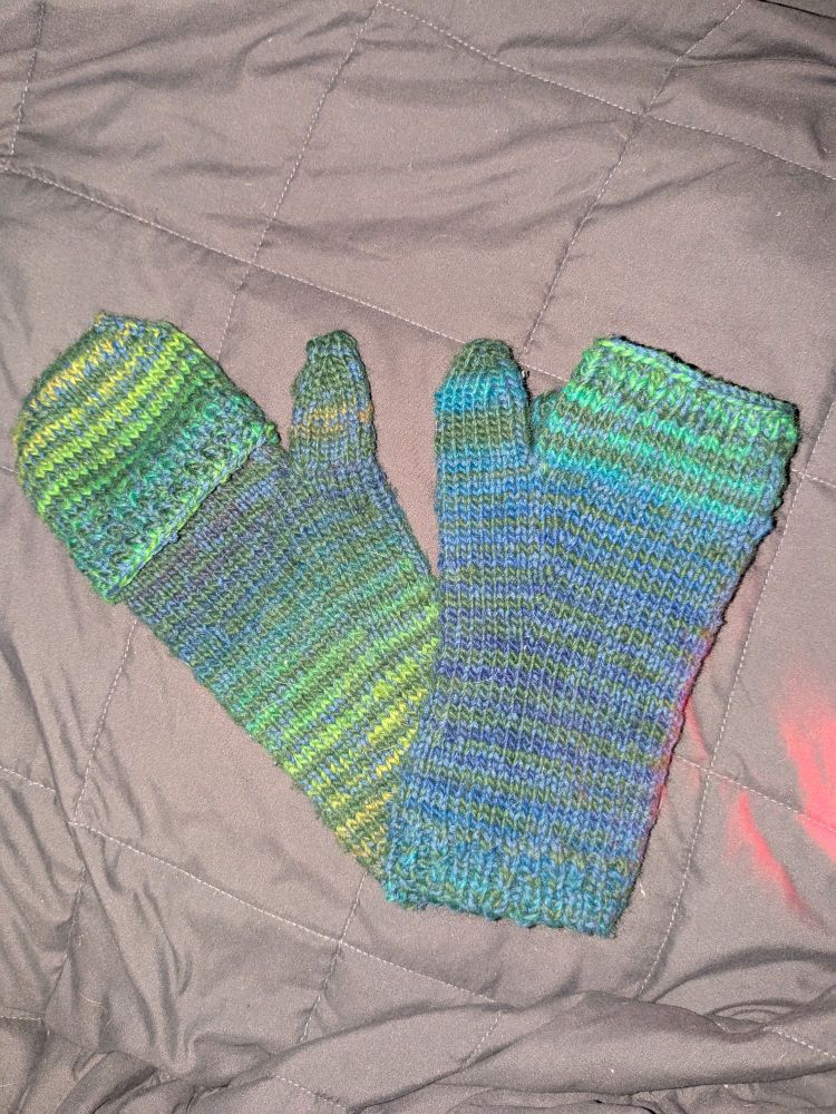 Two mittens in greens and blues on a grey background. One has a cover, one is fingerless 