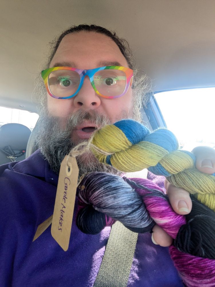 Man in rainbow glasses holding a skein and half a skein of yarn. Full skein (merino/nylon) is in purples and blacks, half skein (merino/angora rabbit) is in yellows and blues.