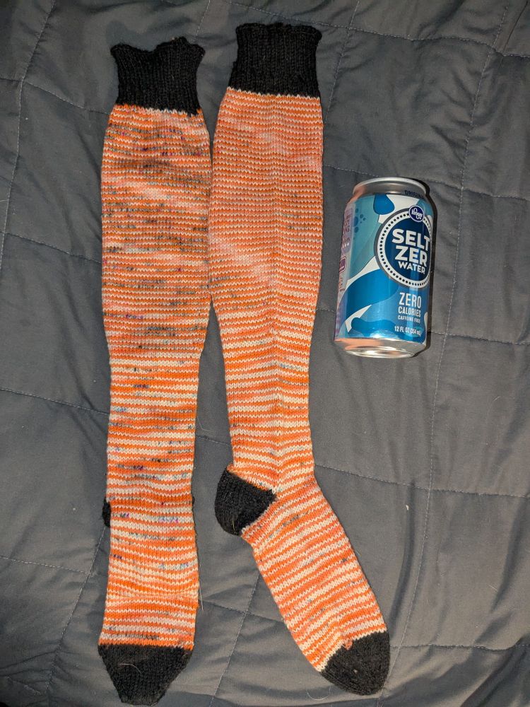 Orange and white striped, speckled socks with black heel, toe and cuff alongside a regular sized can of seltzer water.