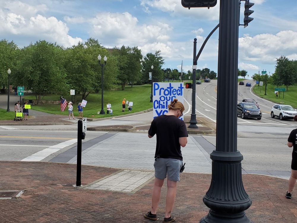 Protesters opposed to Donald Trump hold signs on four corners of a busy intersection in Fairmont, WV. The nearest protestor holds a sign reading 'Protect Our Children - Vote Democrat'.