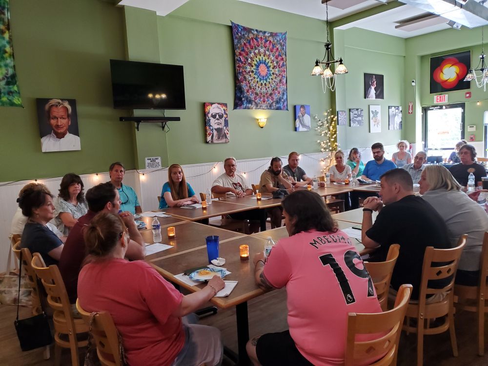 A crowd of people at a restaurant, with multiple tables formed into a circle, talking about local issues - secondary angle.