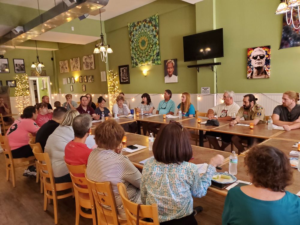 A crowd of people at a restaurant, with multiple tables formed into a circle, talking about local issues.