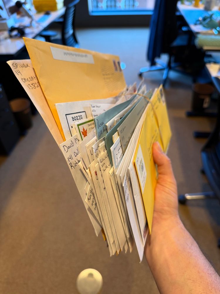 A hand is holding a large stack of assorted envelopes, including yellow and blue ones, with various addresses written on them. The background features a blurred office setting with chairs and tables.
