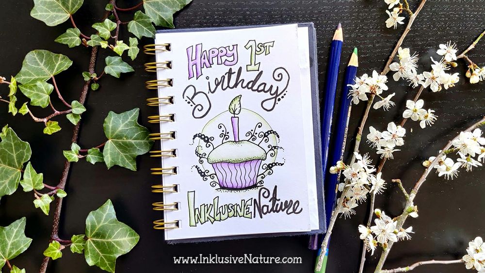 SketchNote reads 'Happy 1st birthday Inklusive Nature'. Photographed in a sketchbook surrounded by pencils, ivy and plum blossom