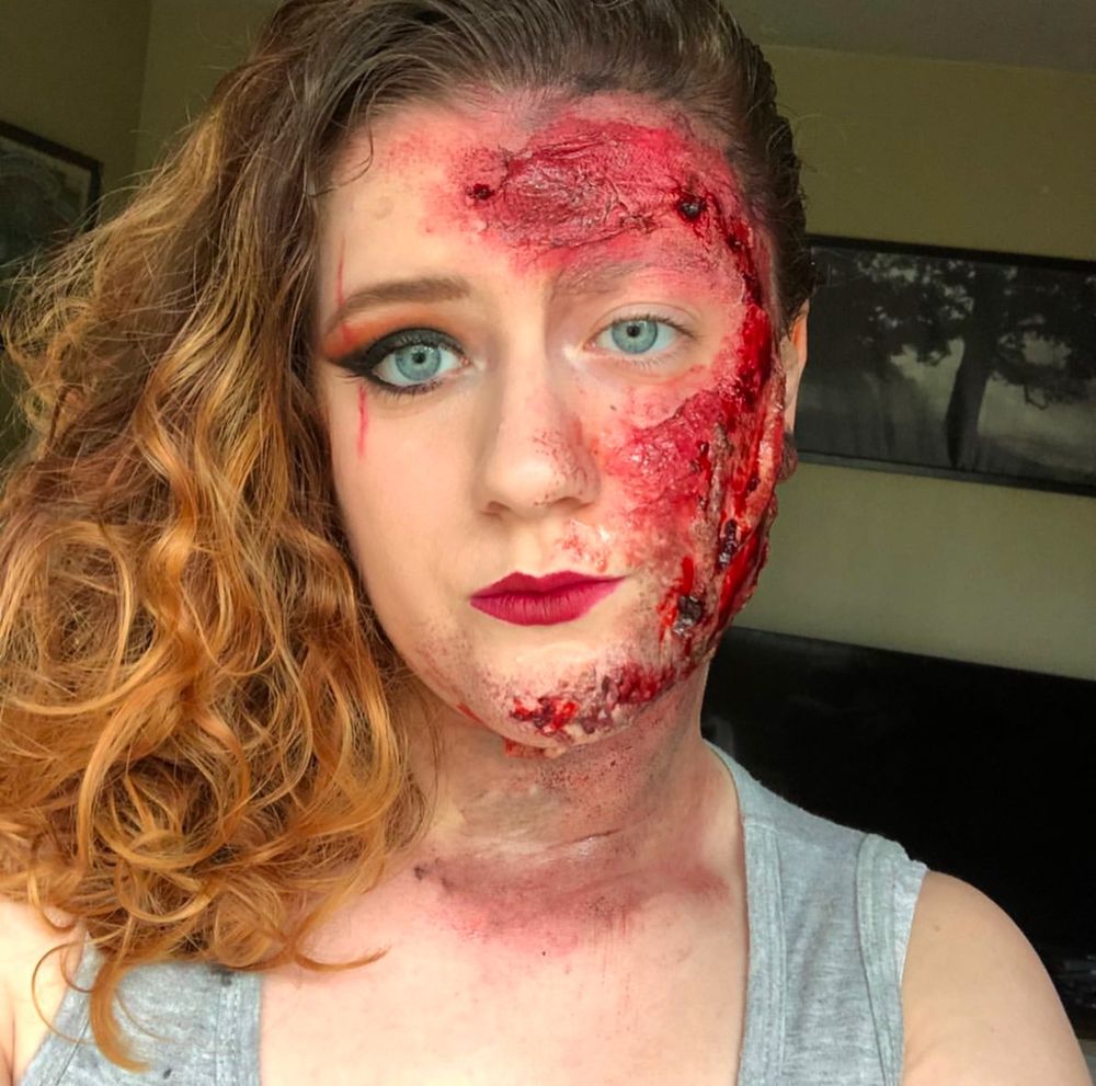 Women with blonde hair looking straight at camera, right half pinned and gelled back. Right side of face has fake burns and left side has red Smokey eye, winged eyeliner and red lips. 