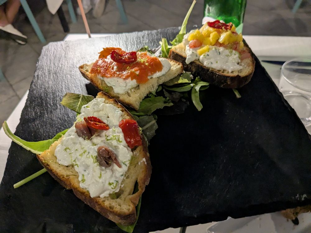 A plate of bruschetta done three different ways, each with burrata, but one has mango, another salmon, another tomato and anchovy