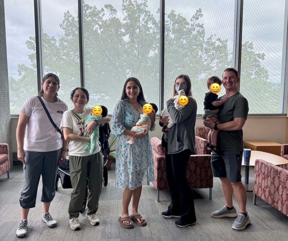 Five smiling adults holding four babies are posing for a photo in front of a large window. Three babies are very small, about 2 months old. The fourth is a toddler. 