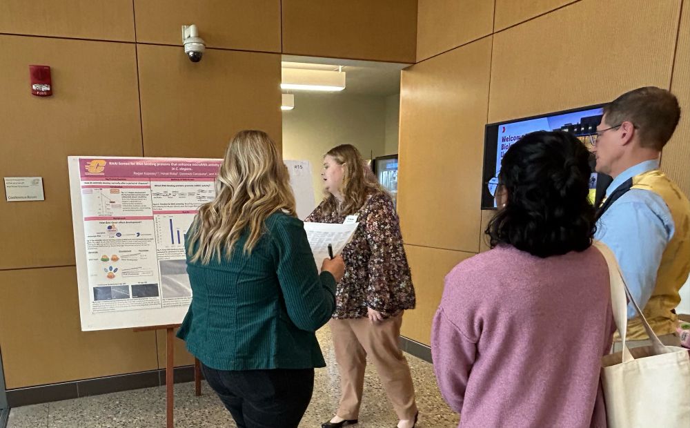 A woman with blond hair and a patterned shirt appears to be pointing at her poster and explaining to the three onlookers. One of the onlookers is centered int the foreground: we see long blond hair and a green jacket. The other two onlookers are on the right. 