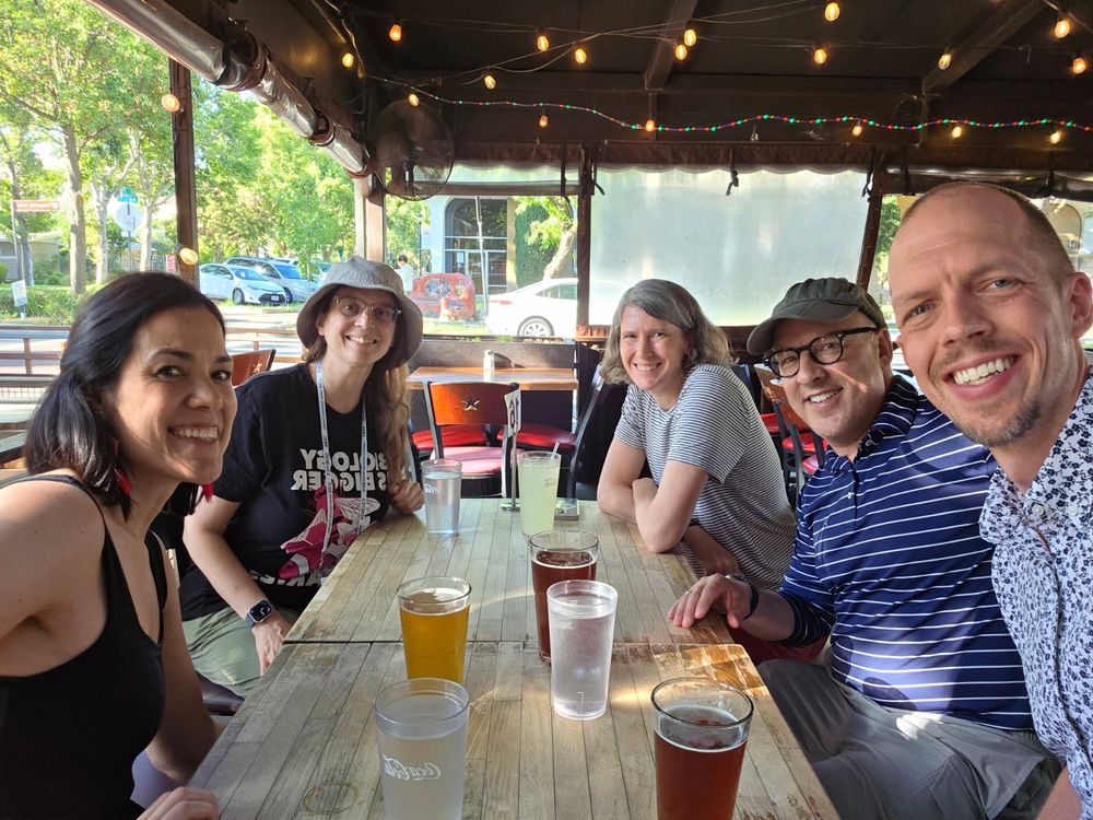 Five people sitting at a wooden table, outdoor restaurant seating, smiling for the camera. Current and former Ambros lab members. 