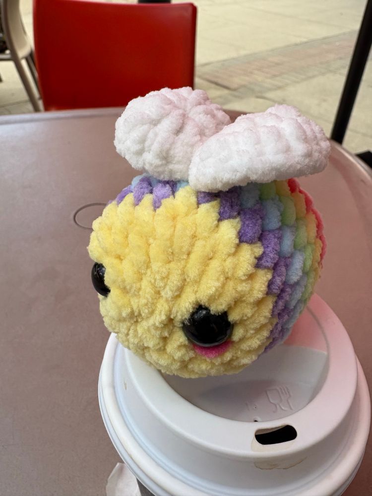 A small crochet bee with rainbow colours on its body sitting on a Togo coffee cup lid 