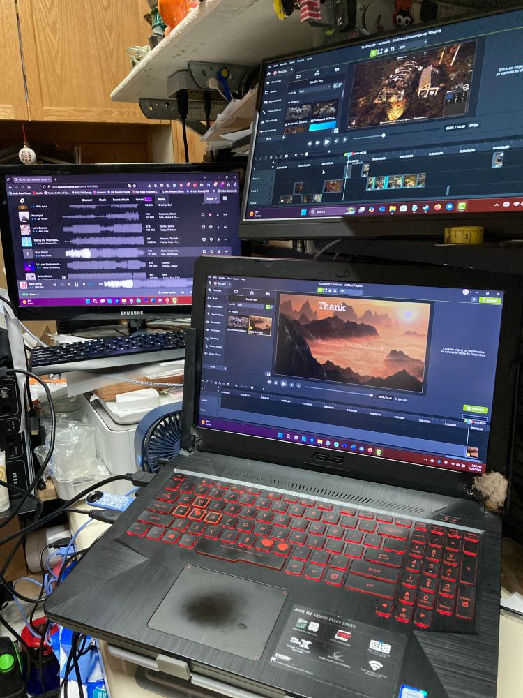 A laptop (with video editing software up) sits among two other computer screens with music (Epidemicsound) and more video editing software tabs.