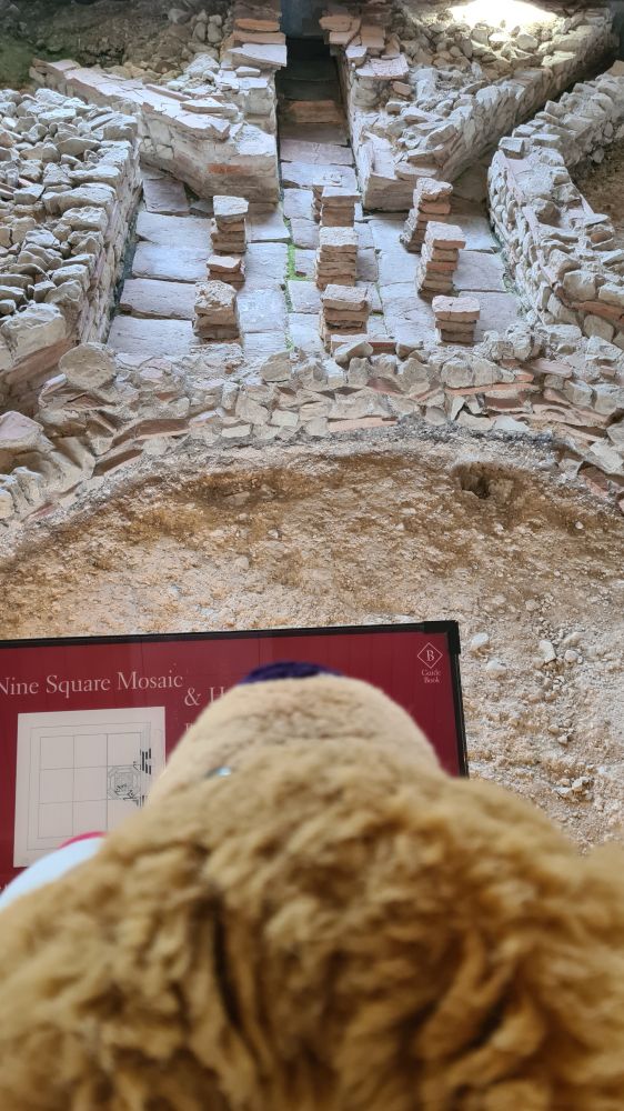 me looking over a lower level and obscuring the sign explaining the hypocaust. so... you have recessed channels in the underfloor where air can pass through, with nine little neat pillars in the middle of a big square recess in the middle of the room. the upper floor is missing but you can see how high it would have been from the filled in stone areas around the recesses. 