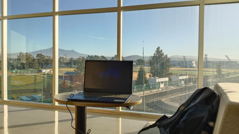 Photo of my laptop on a table with a full view behind through a window. You can see the rest of the uni and some mountains