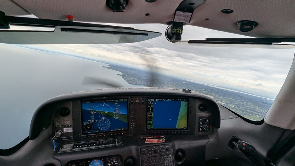 The cockpit of a propeller aircraft banking slightly to the left, flying over a coastline. 