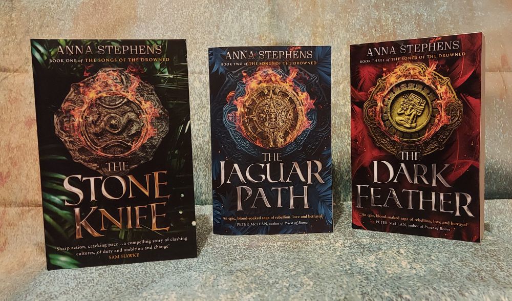 Three books - the stone knife, the jaguar path, the dark feather - stand against a pastel background 