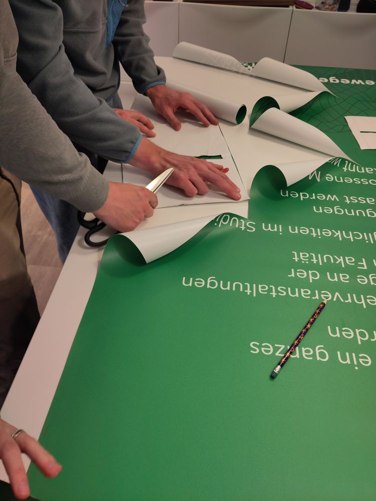 Auf einem Tisch liegt ein Banner mit Bleistift und darauf sieht man zwei Paar Hände, die ein Schnittmuster und eine Schere halten.
