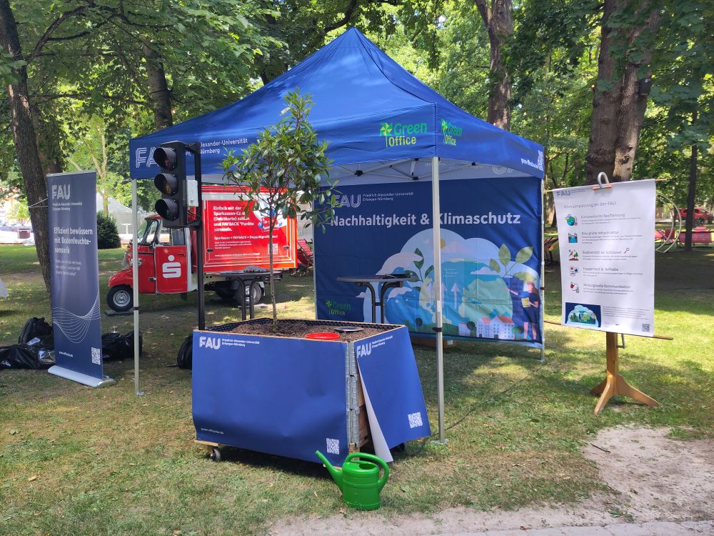 Im Hintergrund ein blauer Pavillon im Vordergrund ein blauer Kübel mit Baum und Ampel, davor eine grüne Gießkanne 