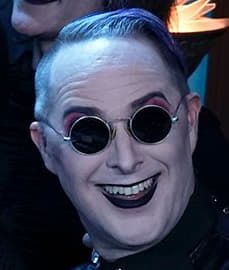 Taliesin Jaffe flashing an unhinged Joker smile in pale makeup and round sunglasses for a Halloween promo shoot