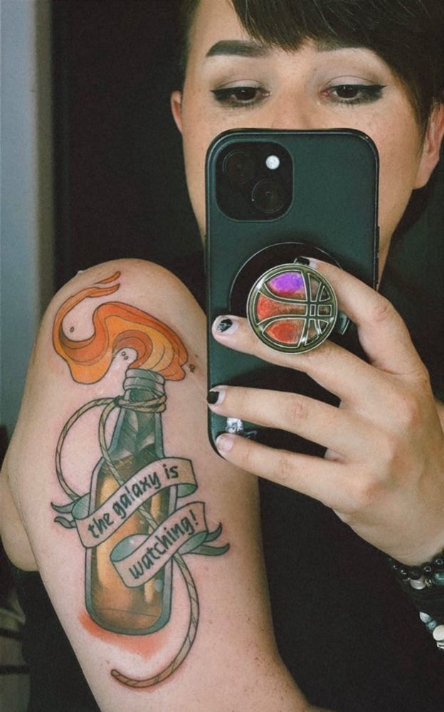 an upper-arm tattoo of a Molotov cocktail with a ribbon that says “the galaxy is watching!” over it