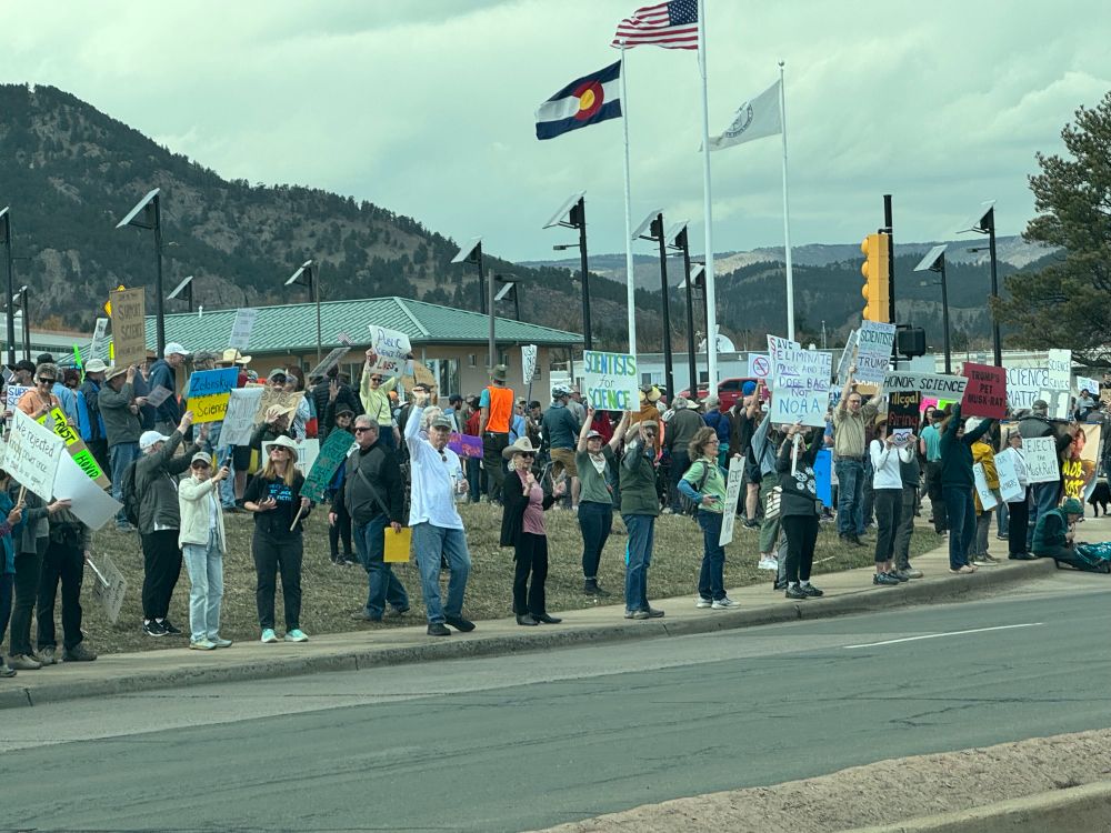 Lots of people with signs rallying by street with US flag, CO flag and maybe a NOAA flag--I can't read it.