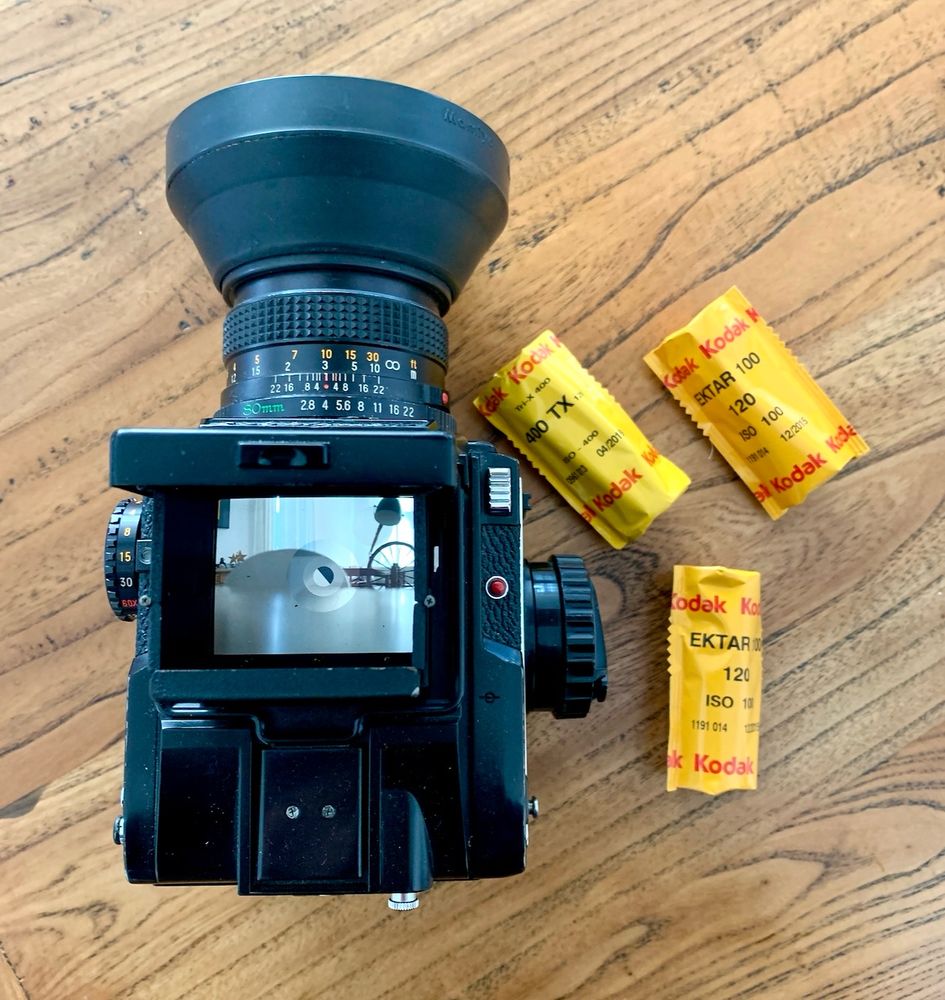 Ein schwarzer Fotoapparat mit Objektiv und Sucher liegt auf einem Holztisch. Daneben befinden sich drei gelbe Kodak-Filmrollen. Der Tisch ist hellbraun, und die dominierenden Farben sind Schwarz, Gelb und Braun. Interessant ist das Bild im Sucher, das ein Raumambiente zeigt.