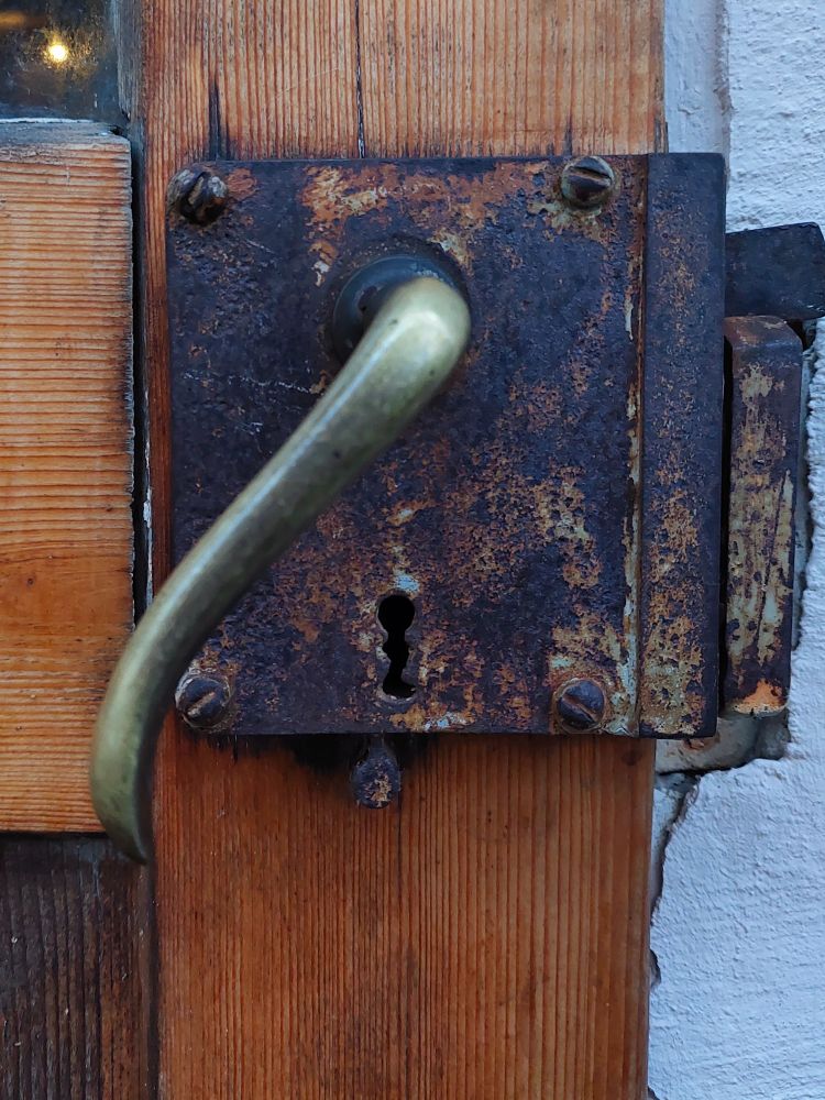 Ein verrostetes Kastenschloß mit herabhängender Türklinke aus Messing, oben zwei Schrauben, unten mittig das Schlüsselloch, montiert auf einer Holztür.