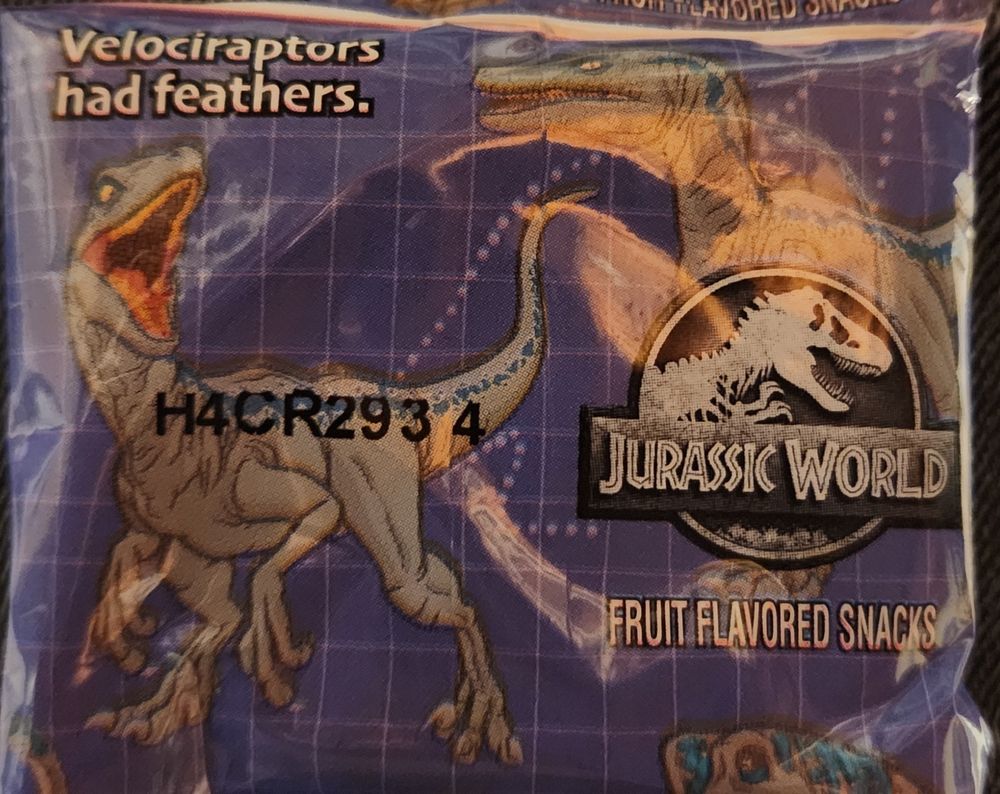 A photograph of a Jurassic World branded fruit snacks package. In the top left it says "Velociraptors had feathers." There are pictures of two Jurassic World style Velociraptors on the package, notably lacking feathers.