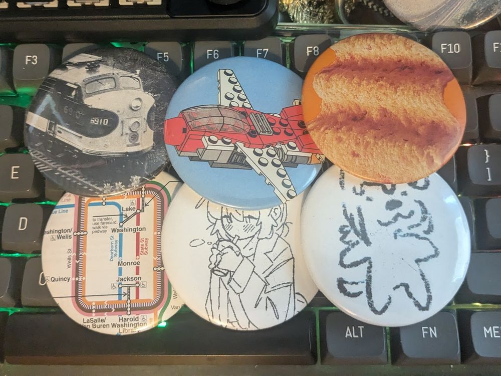 6 buttons laid on top of a keyboard. Left to right a 3/4 view of a F-unit locomotive, a small Lego jet, a close up of a Harvest Cheddar sun chip, a map view of the Chicago L "Loop" tracks, a catgirl drinking a drink, and a crude sketch of a dog-person