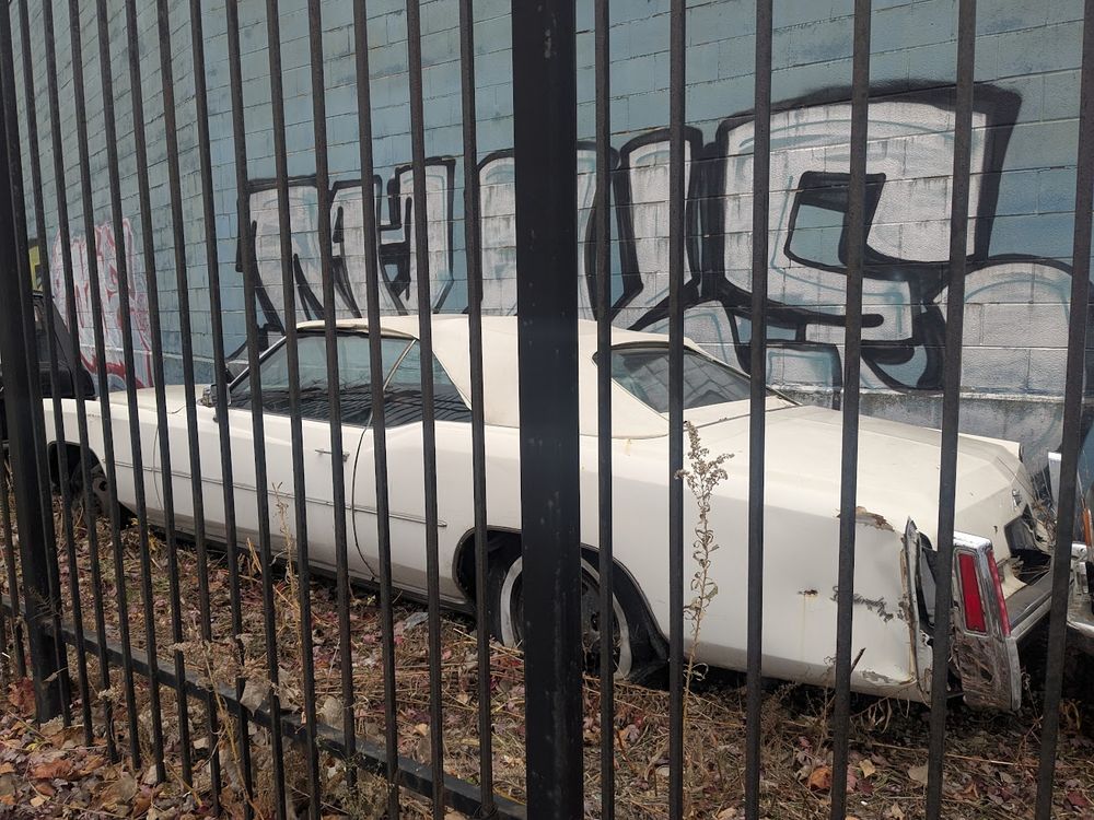 A large, white, "land yacht" Cadillac, sitting behind a fence with severely deflated tires.